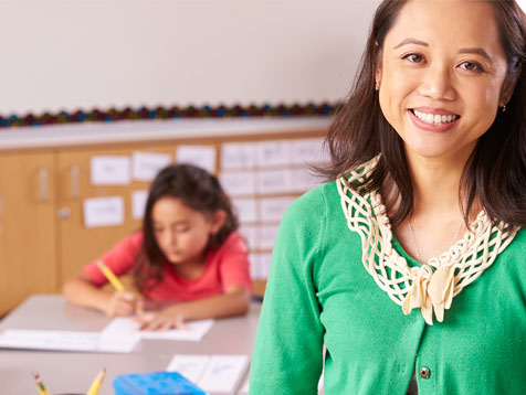 Smiling female teacher in green cardigan in foreground, child student writing in background