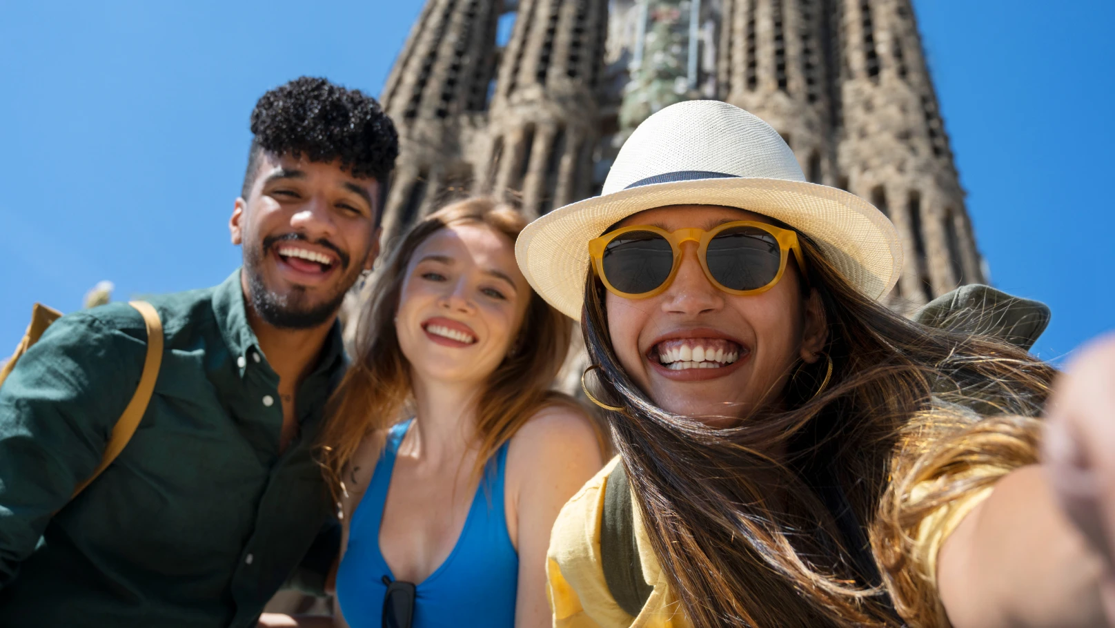 Three people take a selfie in front of the Sagrada Familia in Barcelona, with the basilica’s ornate towers rising behind them under a clear blue sky