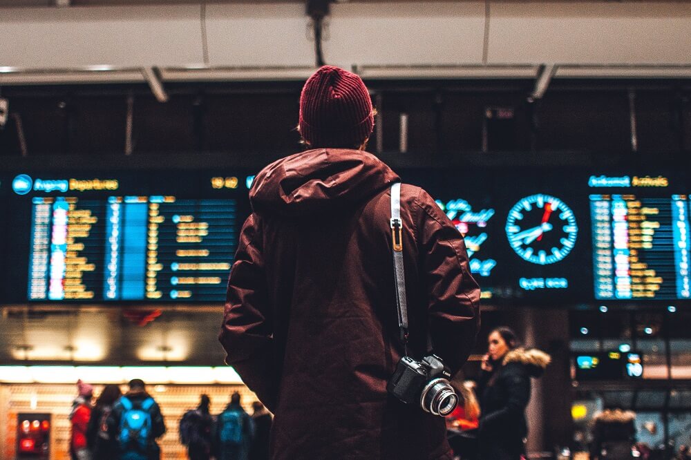 Male in red beanie and red jacket with camera over right shoulder looking up at departures board