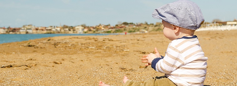 Clothed male baby wearing a flat cap sat on a beach with sand in his hands