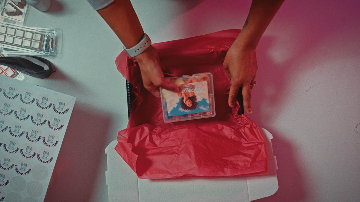 A person packing a small plastic container with an image of a woman into a box lined with red tissue paper, with stickers and packaging materials nearby