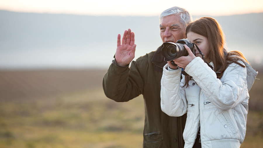 Father teaching his daughter how to use a camera surrounded by afternoon sunlit green grassy hills