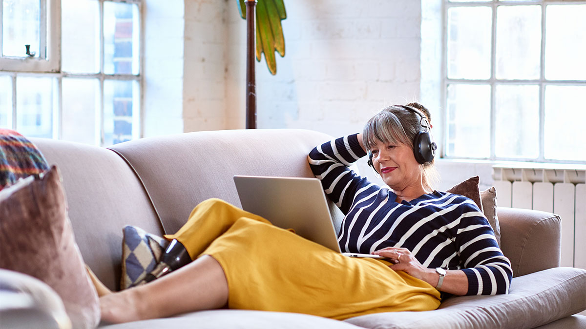Woman wearing headphones who laying on a couch looking at the screen of a laptop on her lap