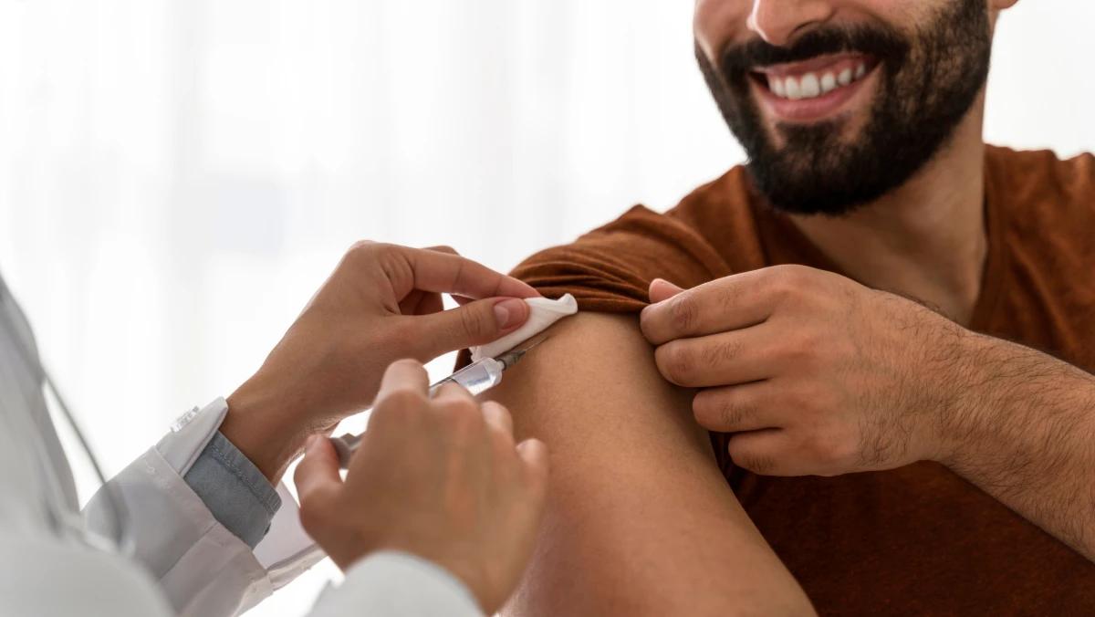 A healthcare professional in a white coat gives an injection in the upper arm of a person wearing a brown shirt, using gauze for support