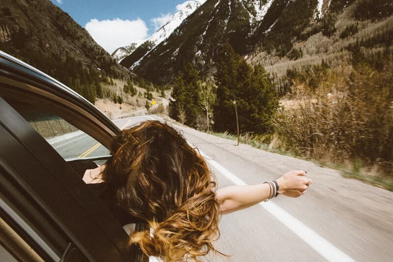 Car driving down winding mountainous road with woman leaning her head and arm out of passenger window