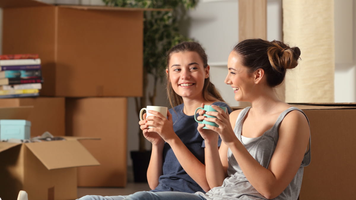 Two girls laughing sitting on the floor holding a mug with cardboard boxes in the background