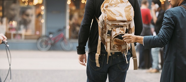 Man with rolled sleeve taking item from backpack on another man walking in ahead in a street