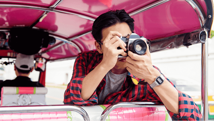 A male passenger in a pink tuk-tuk leans out the back, holding a camera, taking photographs