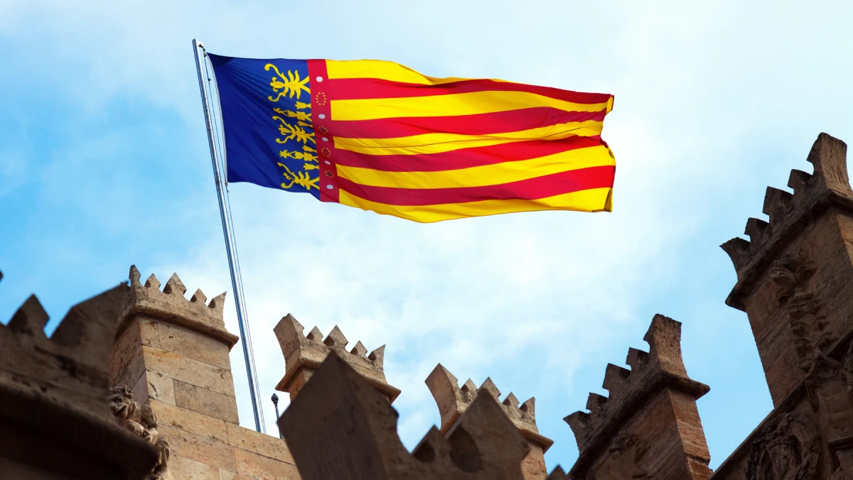A Valencia flag with yellow and red stripes and four yellow lions on blue flies above a stone building with crenelated battlements