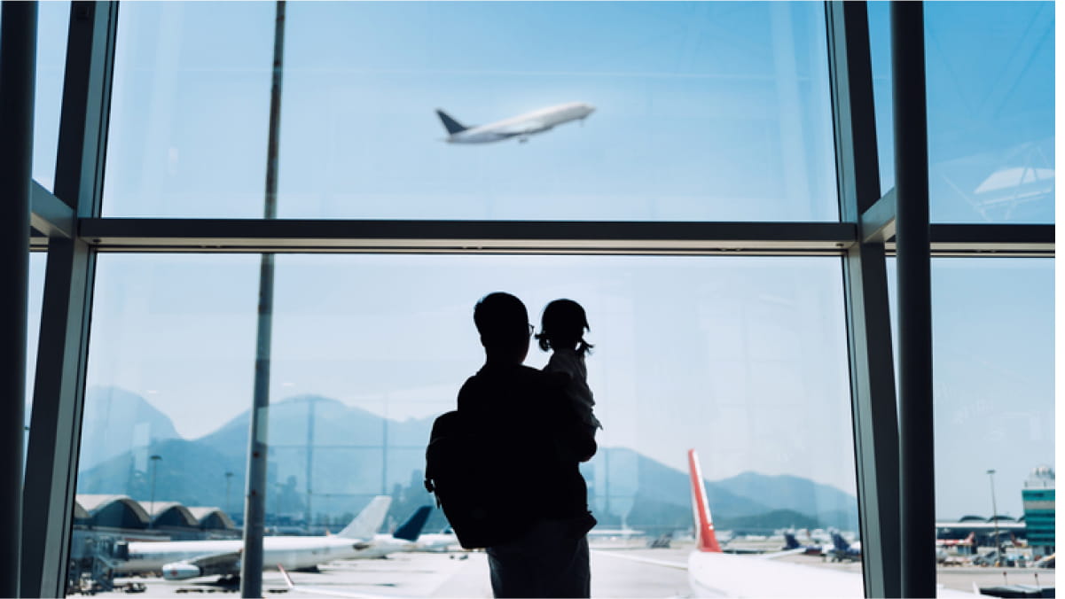 An adult holding their child looking out of the airport window at a plane taking off in the distance