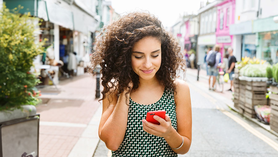 Young woman in a high street walking towards us while looking down at her phone in her left hand