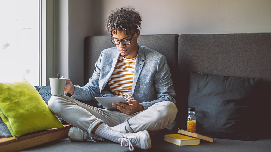 Man sitting with legs crossed looking at his tablet in one hand and a cup of drink in another hand