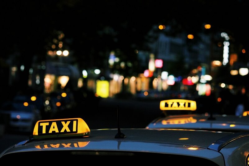 Illuminated taxi signs on taxi vehicles in foreground, late evening street and lights in background