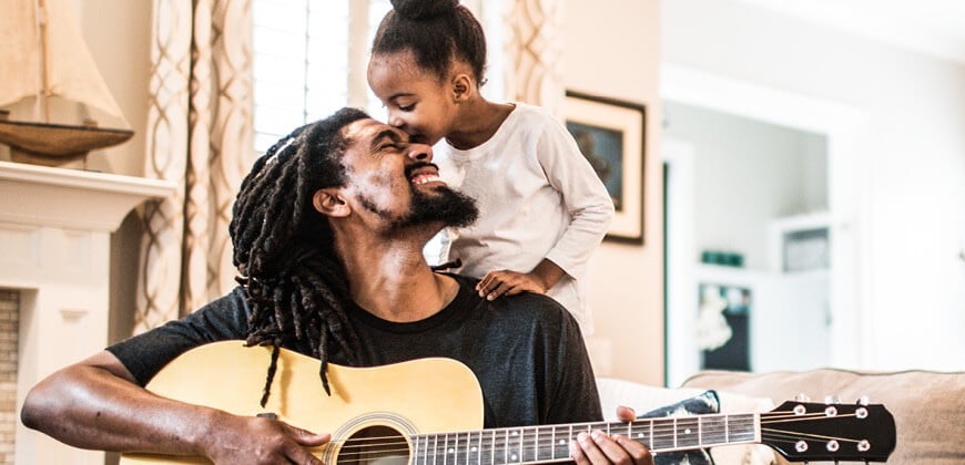 Little girl kissing her daddy on the forehead while he's playing a guitar, in their home
