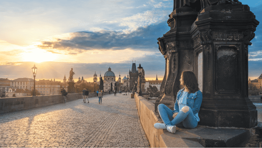 A woman in denim sat on the wall of a bridge in Hungary, watching people and sun set in the distance