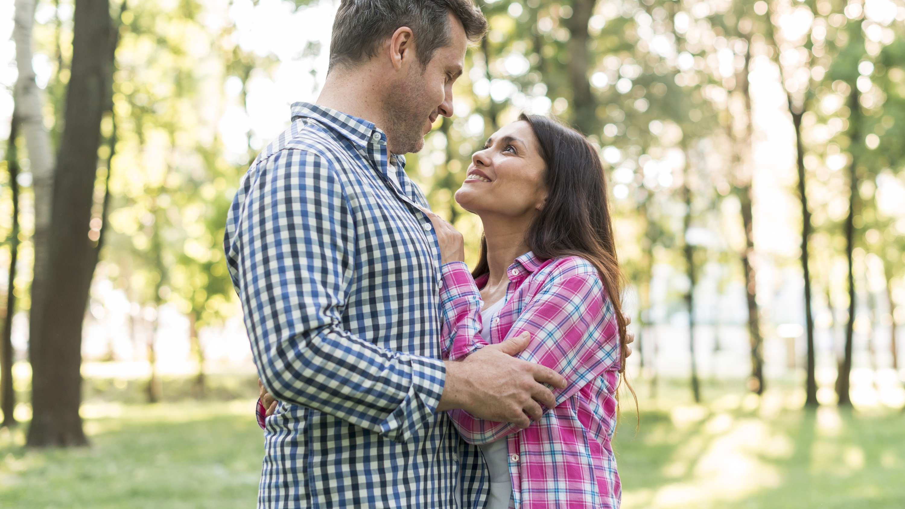 Two people stand close together outdoors, with trees and sunlight in the background