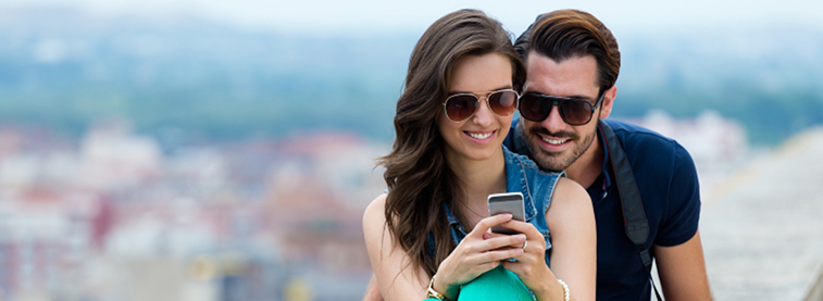A couple wearing sunglasses and smiling while looking at something on the woman's phone in her hands