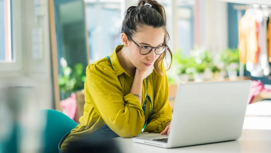 Young woman sat at a table wearing glasses looking at her laptop