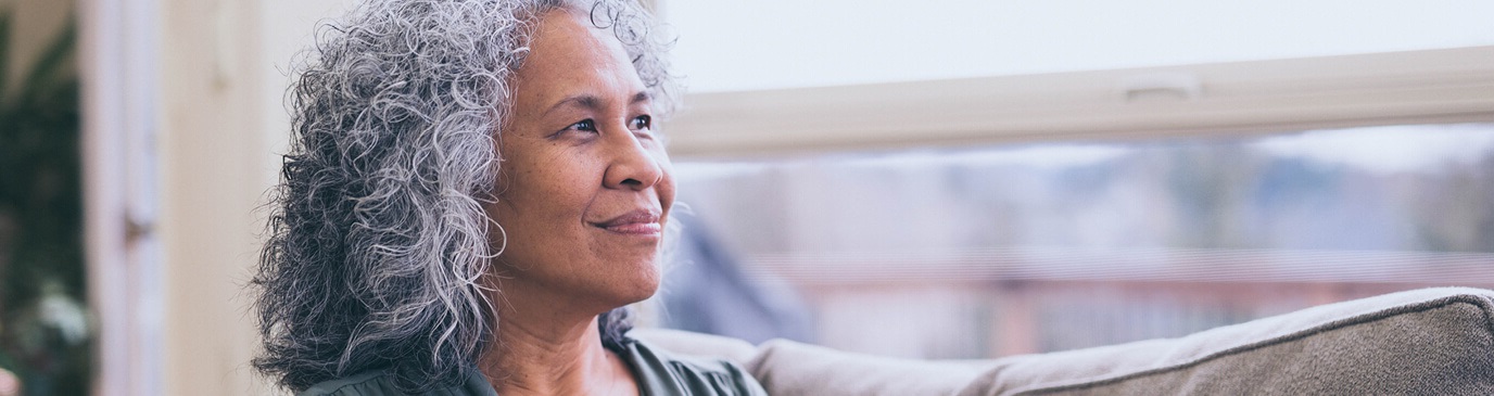 Mature woman sat on a couch, smiling as she looks out of the nearby window