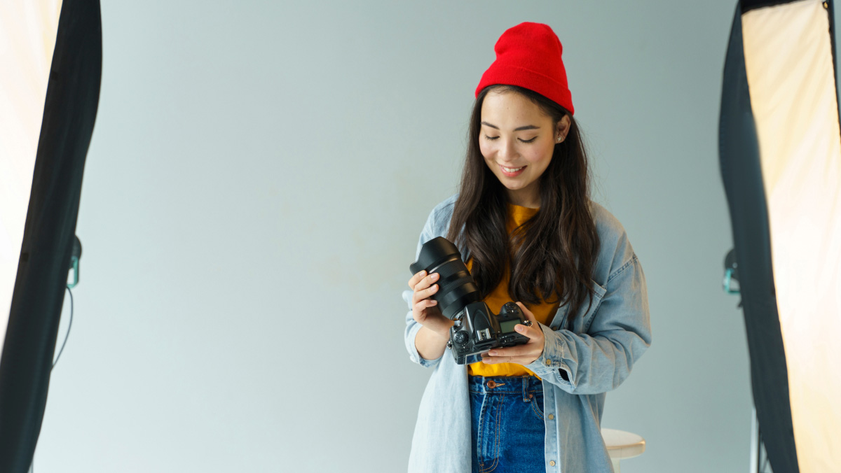 A person in a red beanie and denim jacket stands in a photo studio, holding a camera between two big softbox lights