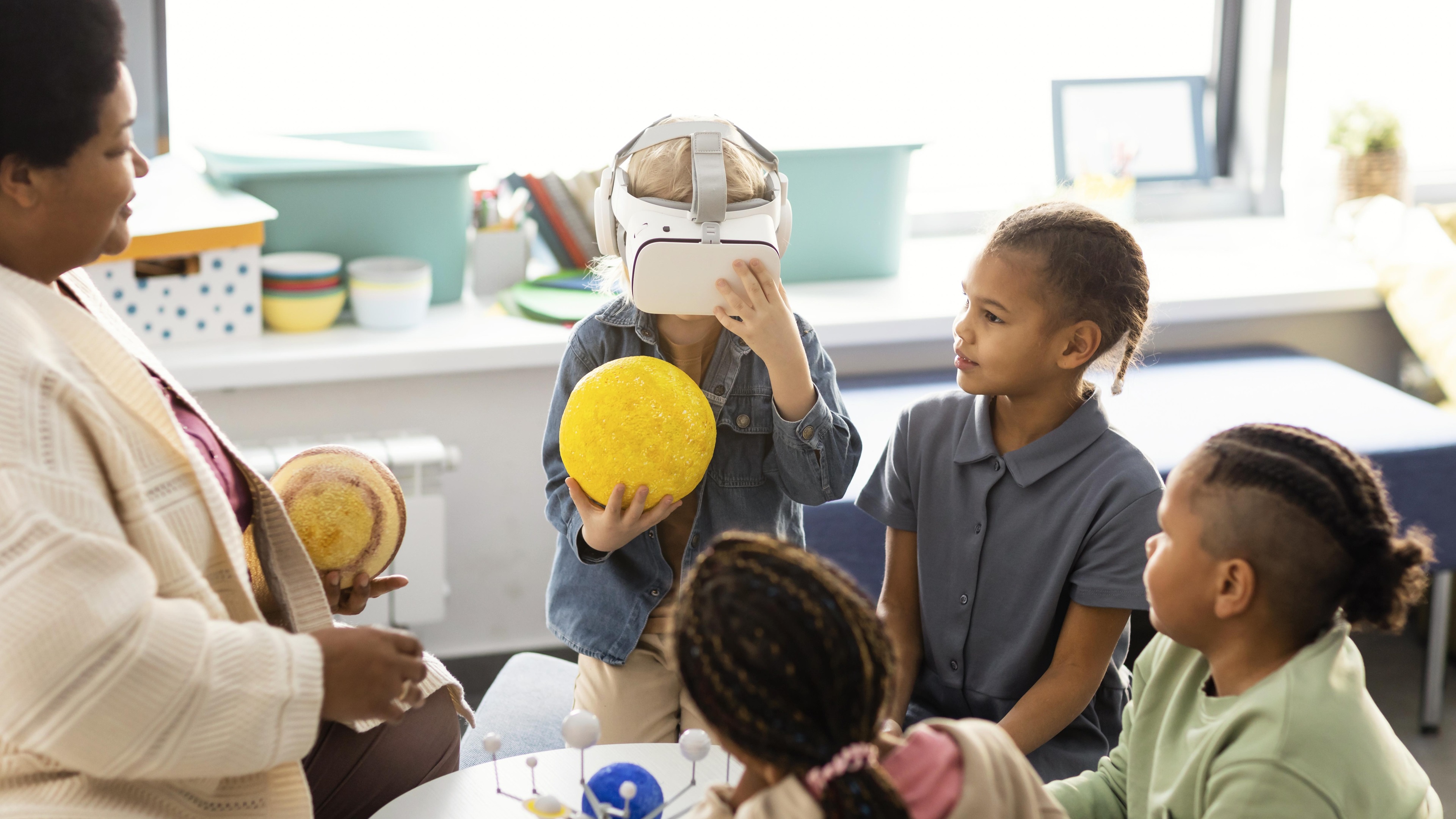 A child wearing a VR headset holds a yellow sphere resembling the sun. Three other children and an adult look on in a nursery or classroom setting