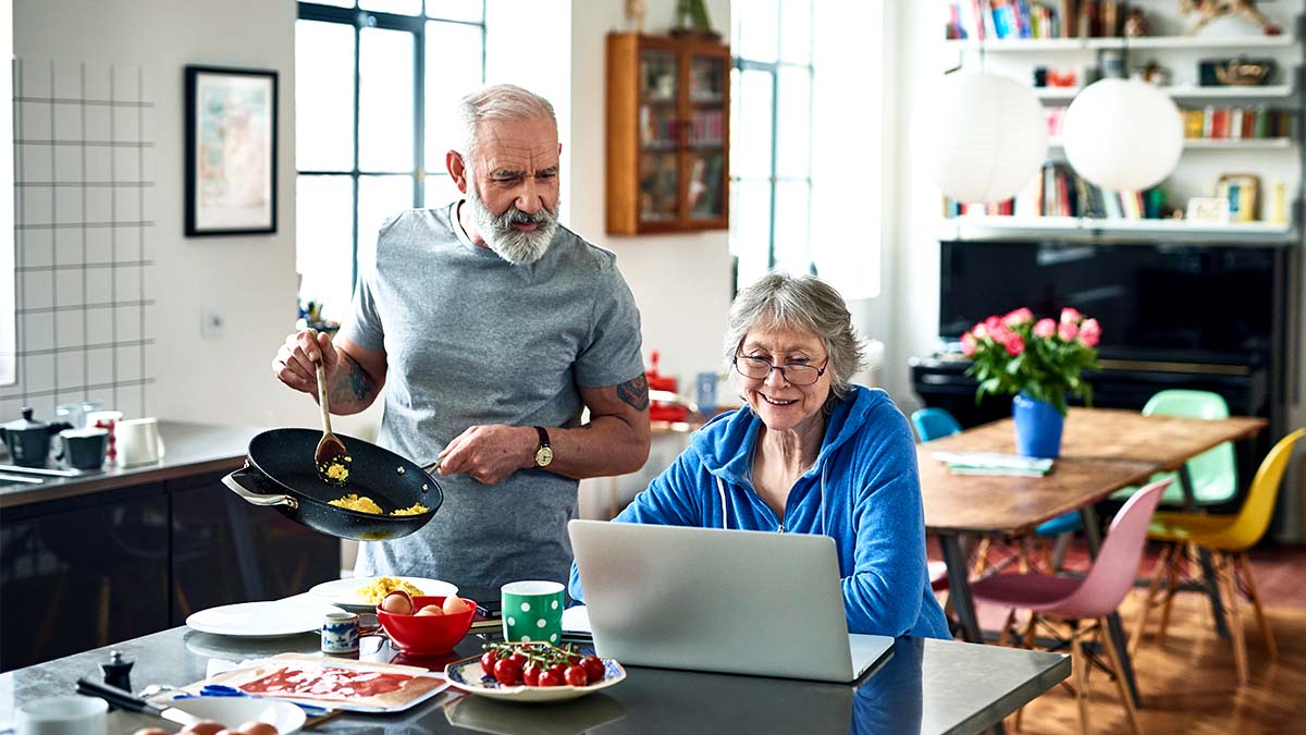 Elderly couple in the kitchen looking at the laptop screen, while man is cooking food holding a pan and spoon