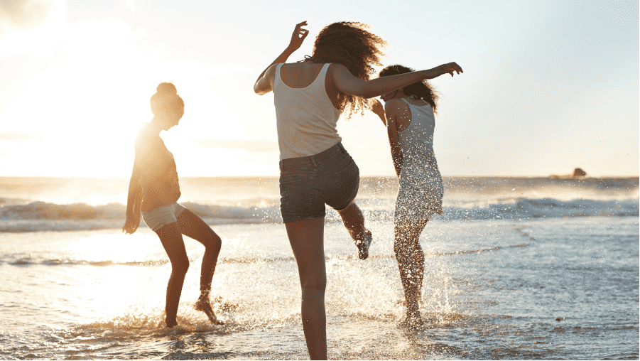 Three young women on a beach jumping and kicking the shallow waters at each other as the sun sets