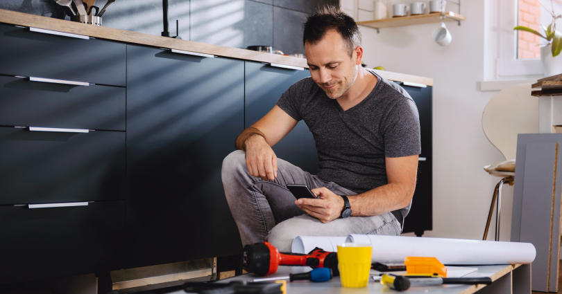 Man kneeling down on one knee looking at his phone with DIY tools in front of him