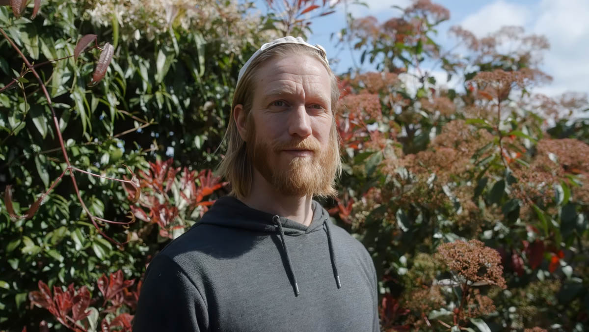 A bearded man in his thirties stands outdoors on a sunny day, his long hair pushed back by a hairband. He looks warmly towards the camera 