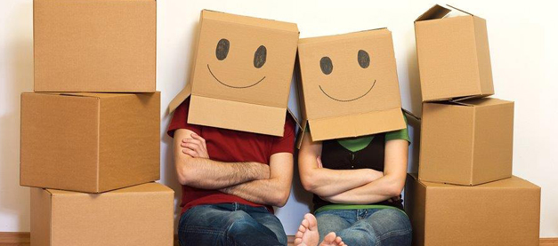 Couple sat between stacked boxes, both wearing boxes on their heads with smiley faces drawn on them