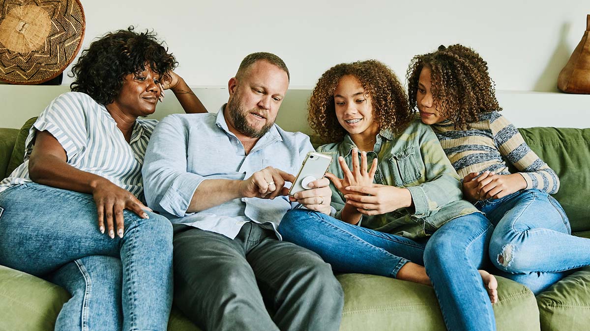 A family of four sitting on the sofa, all looking at the mobile phone held by the father