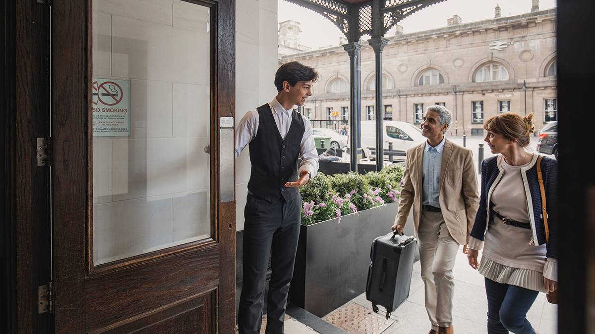 Hotel staff member holds door open as two guests approach with luggage