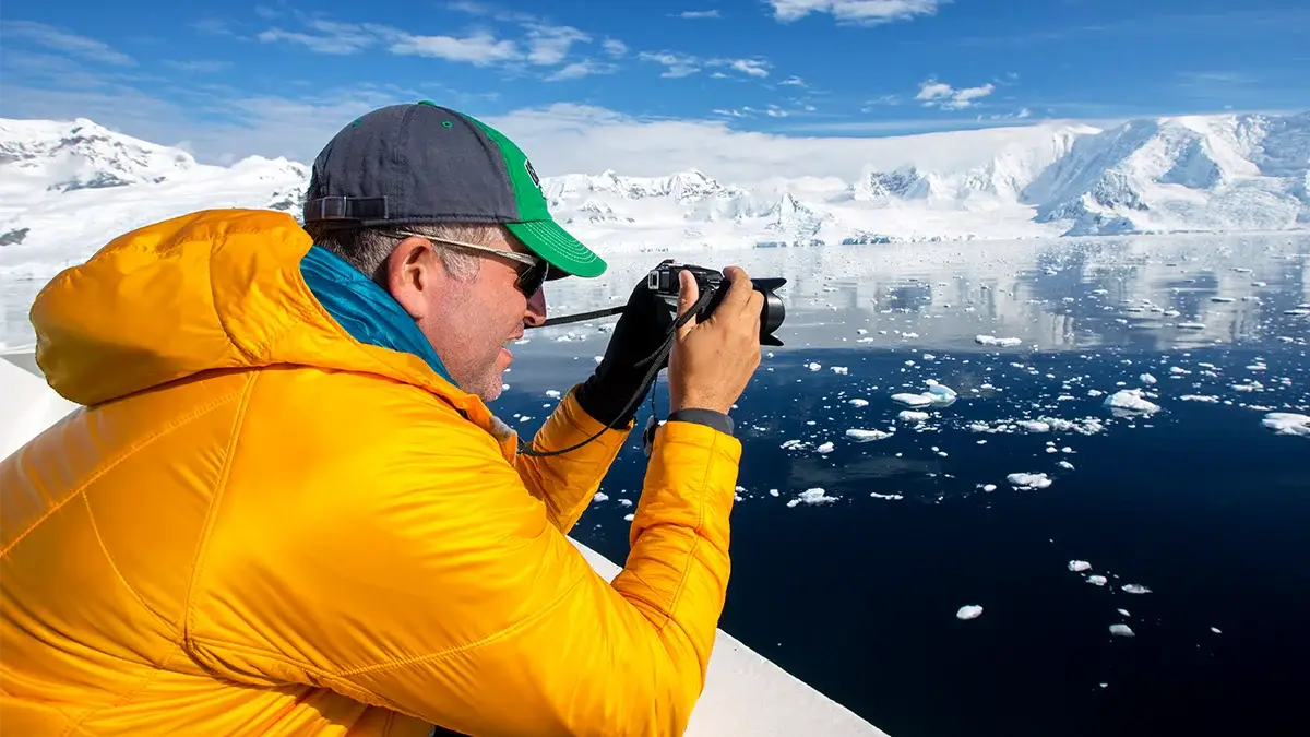 Man in an arctic scene wearing a yellow winter and cap, holding a camera up taking a picture