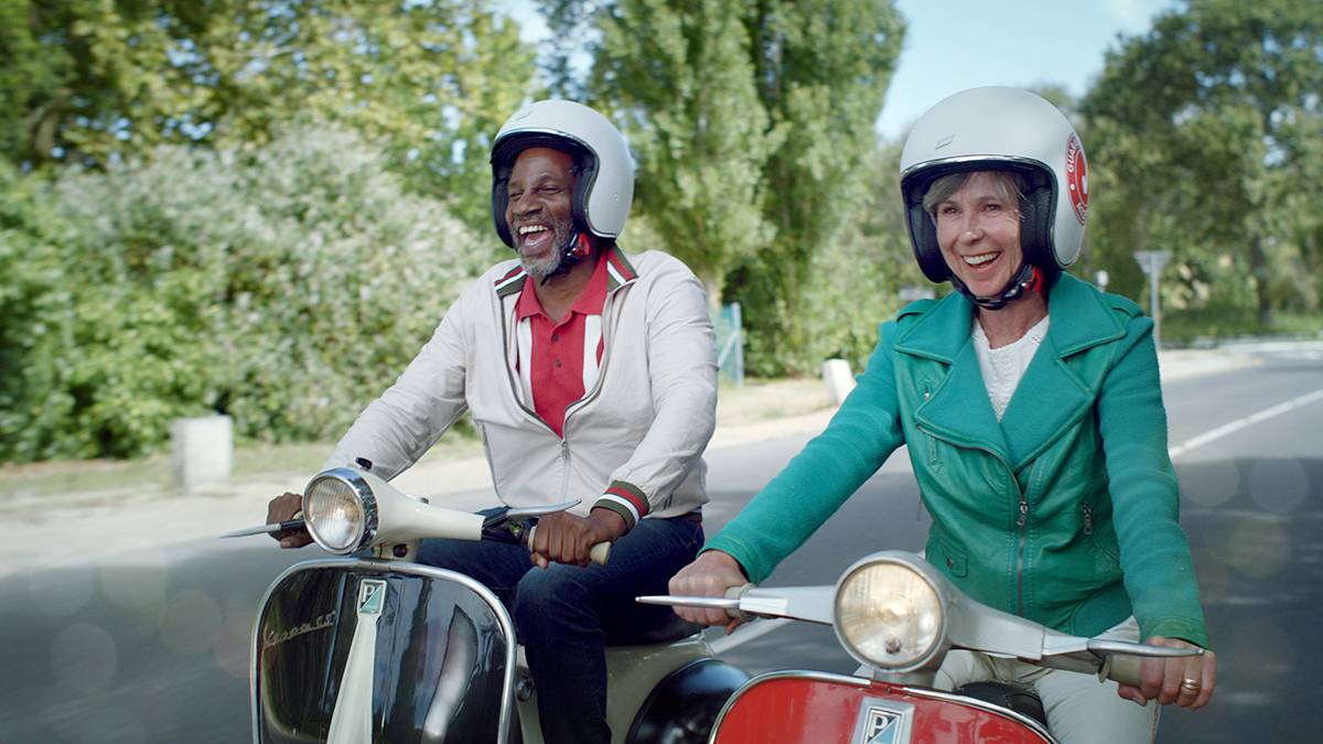 Two people wearing helmets ride scooters side by side on a scenic road lined with trees