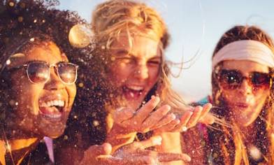 Three young women, left and right both wearing sunglasses, all laughing in the sunshine
