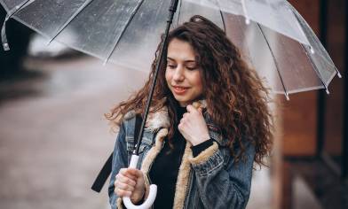 Person holding a transparent umbrella outdoors, wearing a denim jacket. The background shows a wet pavement and buildings