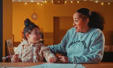 A child holding a pencil sits at a table with an adult wearing a 'SMELL-A-LICIOUS' sweatshirt