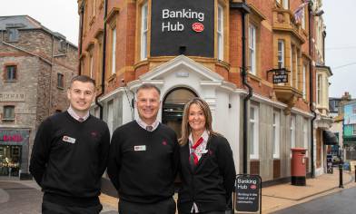 Three people in black uniforms stand outside a red brick building labelled 'Banking Hub' with white trim and large windows. It proudly stands on a high street