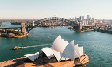 Sydney opera house in the foreground with Sydney Harbour Bridge beyond and a city skyline further back still. A ferry crosses the water between the opera house and the bridge