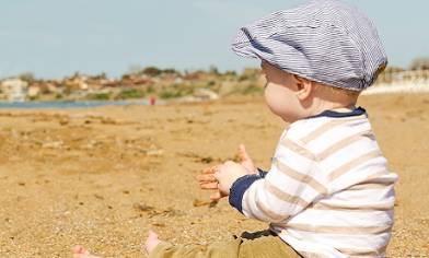 Clothed male baby wearing a flat cap sat on a beach with sand in his hands
