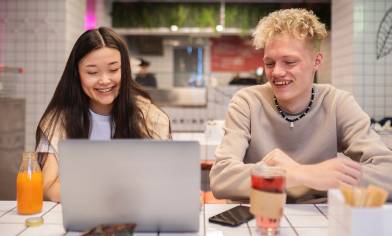 Two smiling people are hanging out at a café table with snacks and drinks, looking at a laptop. The surrounding walls and tabletop are made of white tiles