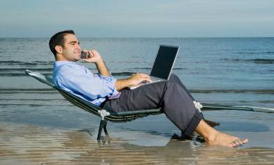 man wearing trousers and a button down shirt with his laptop and pone lounging on a beach