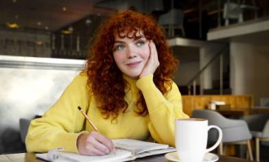 A person writing in a notebook with a warm drink on the table beside them. 