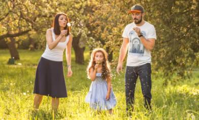 A family stands in a sunlit meadow blowing dandelion seeds surrounded by tall grass and trees