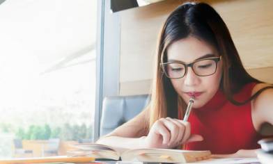 Woman sat in a well-lit café, holding pen to her mouth while reading from a book for her studies