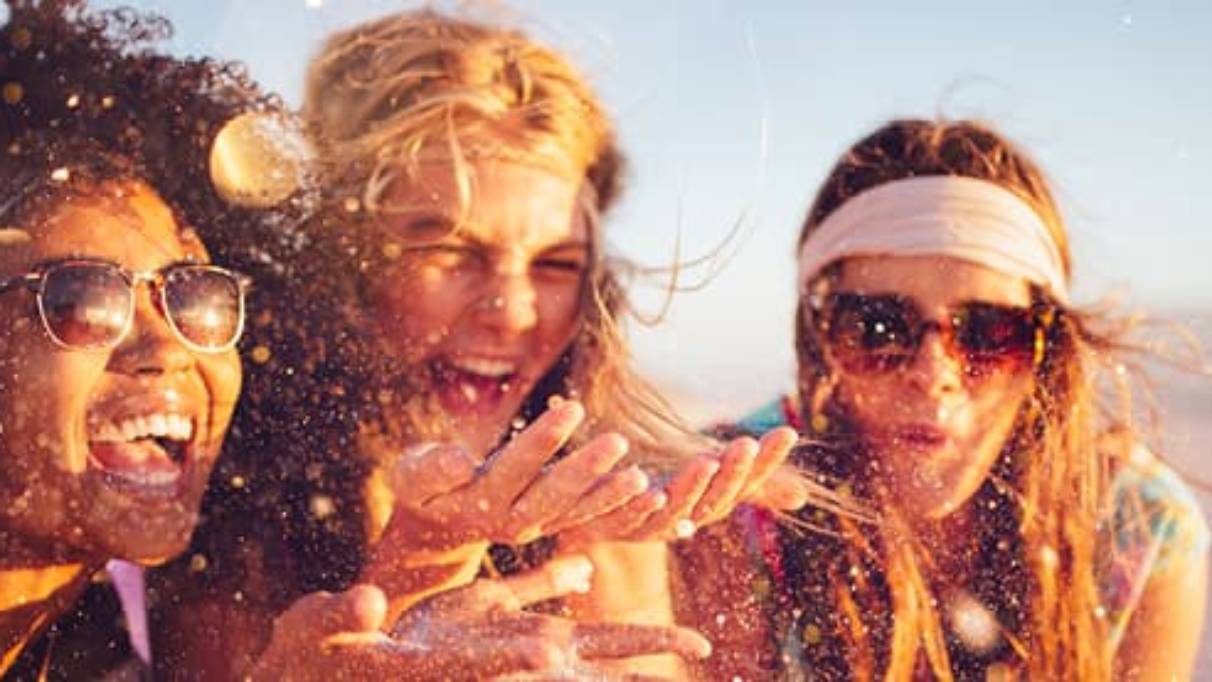Three young women, left and right both wearing sunglasses, all laughing in the sunshine