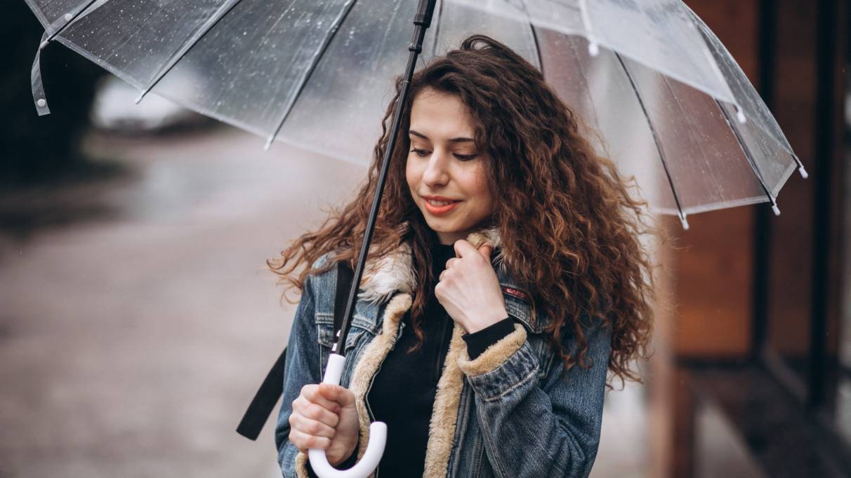 Person holding a transparent umbrella outdoors, wearing a denim jacket. The background shows a wet pavement and buildings