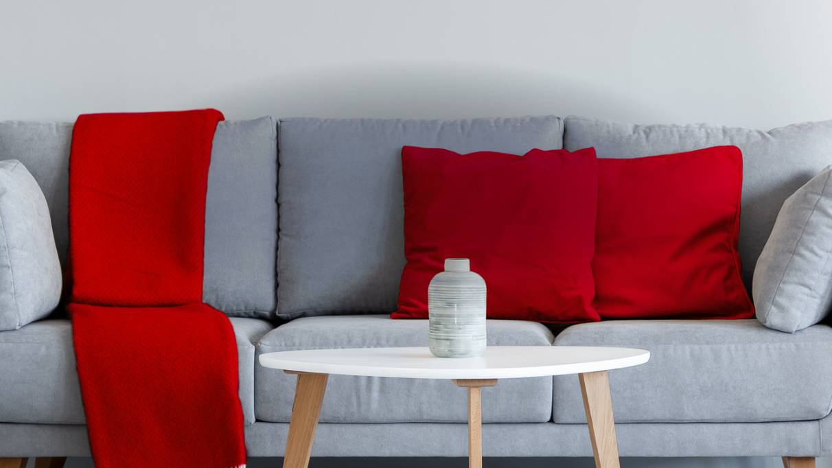Lounge area with plants on cabinets either side of a grey sofa with red blanket, red and grey cushions on it with a table in front of the sofa