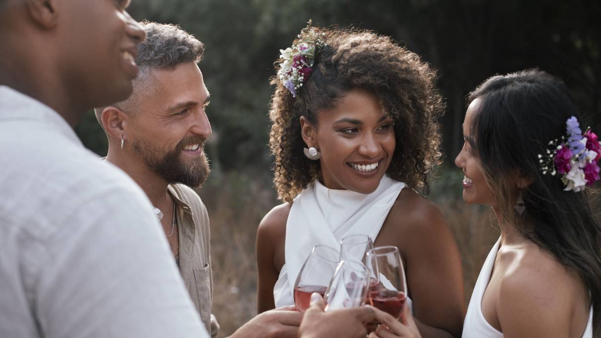 People dressed in light-coloured outfits clinking glasses of wine outdoors, with floral hair accessories visible
