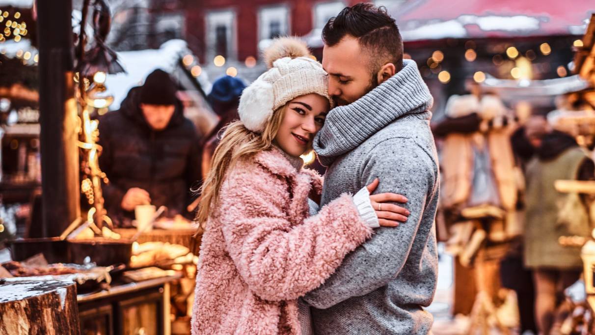 A couple hugging in wintery clothes at an outdoor Christmas market, surrounded by stalls and vendors